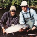 Two men holding a large fish while standing on the bank of a river.