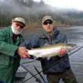 Two men holding a fish while standing on the shore.